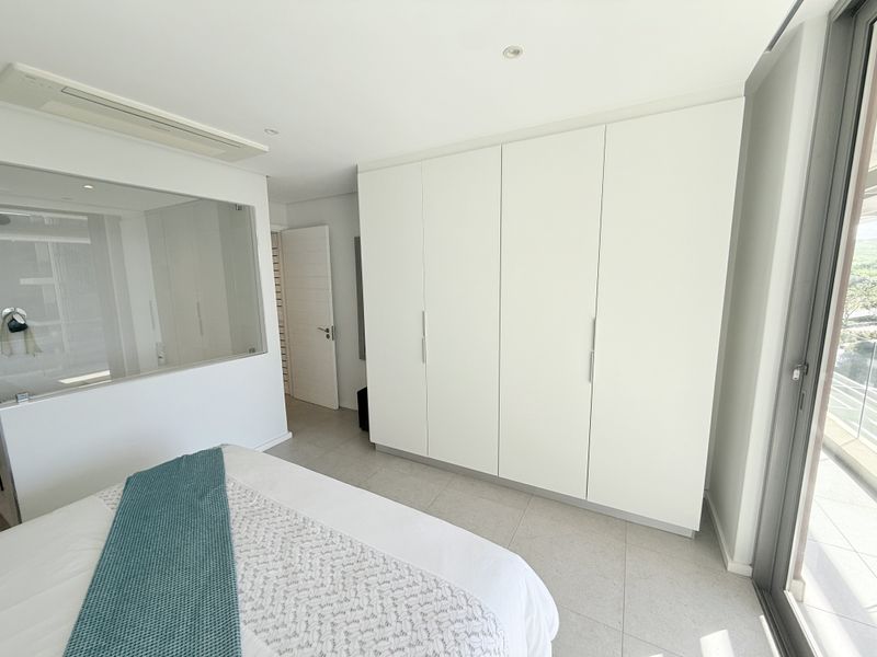 Modern bedroom featuring sleek built-in wardrobes, tiled flooring, and a glass partition wall looking into the en-suite bathroom, with access to a private balcony.