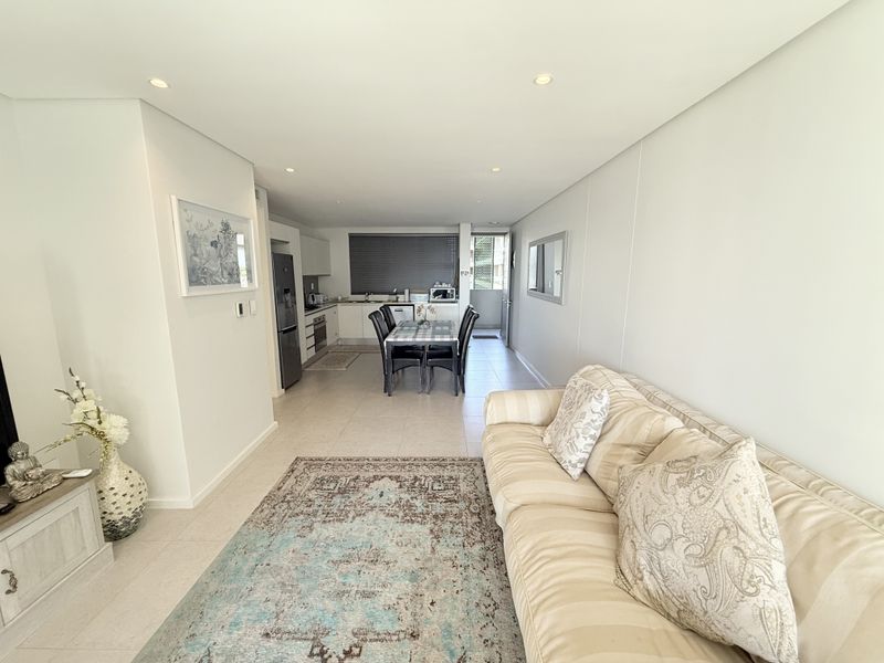 A bright open-plan living and dining area featuring tiled floors, recessed lighting, and a modern kitchen with built-in cabinetry and integrated appliances.