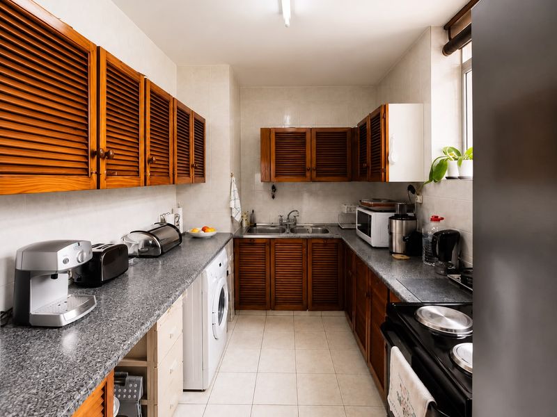 A functional galley-style kitchen featuring louvered wooden cabinetry, laminate countertops, a double stainless steel sink, and integrated laundry plumbing.