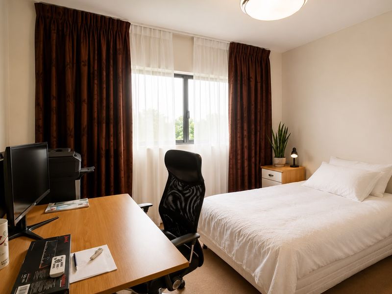 A well-lit bedroom featuring neutral walls, carpeted flooring, and a large window with curtains, providing a comfortable and functional living space.