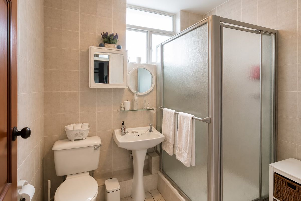 A functional bathroom featuring a pedestal sink, a shower enclosure with frosted glass doors, and neutral ceramic wall and floor tiling.