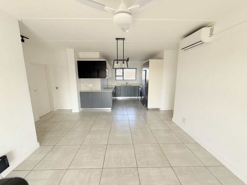 An open-plan living and kitchen area featuring modern grey cabinetry, tiled flooring, a ceiling fan, and a wall-mounted air conditioning unit.
