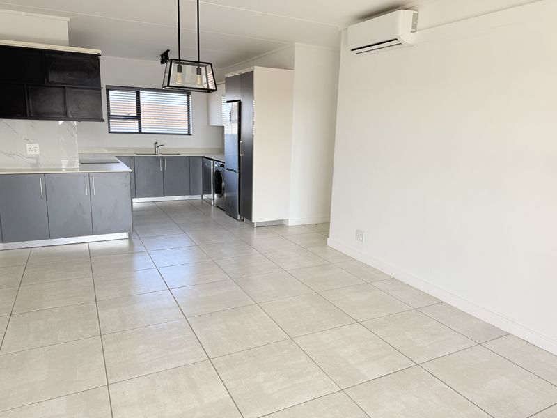 A bright, open-plan living and kitchen area featuring modern grey cabinetry, stone countertops, tiled flooring, and a wall-mounted air conditioning unit.