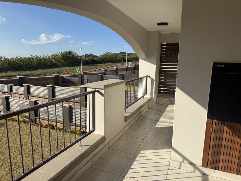 A spacious covered balcony featuring tiled flooring, a built-in braai, and a metal balustrade, offering views of the surrounding estate.
