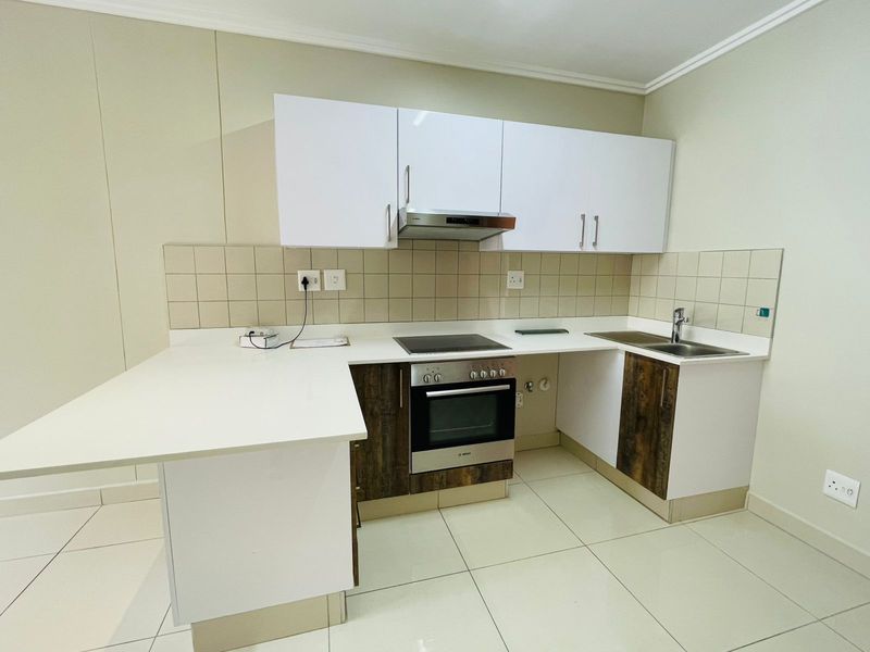 A clean, modern kitchen featuring white and wood-grain cabinetry, stone countertops, a built-in oven, electric hob, and a stainless steel sink.