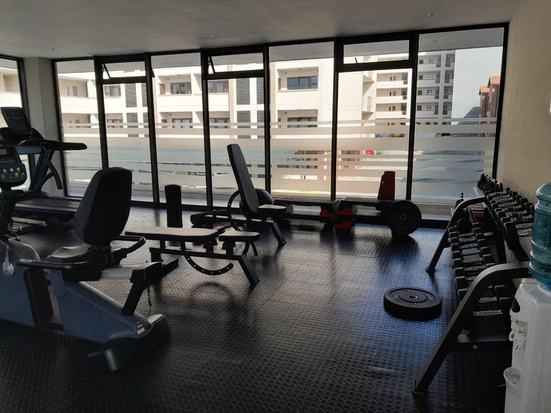 A well-lit apartment gym featuring durable rubber flooring and expansive floor-to-ceiling windows providing natural light and views of the surrounding complex.