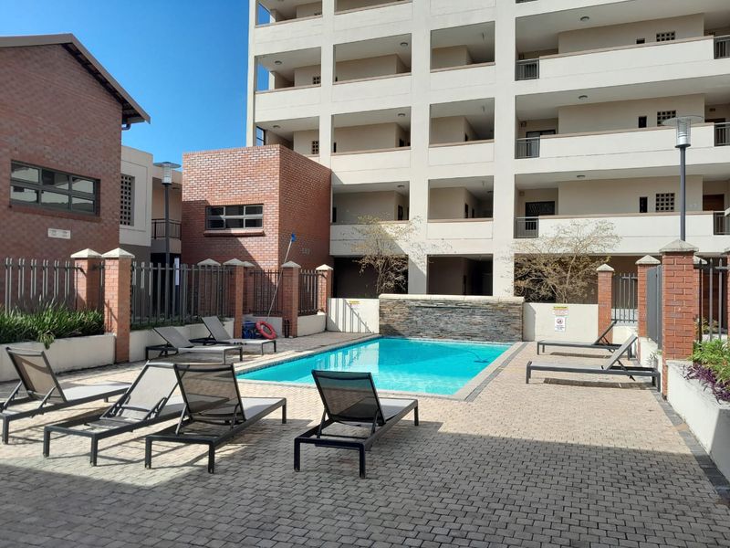 A well-maintained communal swimming pool area featuring a stone water feature, paved decking, and secure brick and metal perimeter fencing within an apartment complex.
