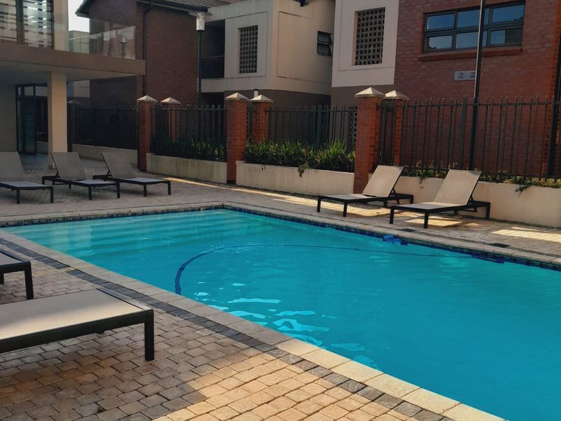 A well-maintained communal swimming pool area featuring paved decking, perimeter fencing, and brick architectural detailing within an apartment complex.