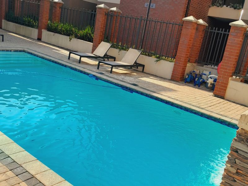A well-maintained communal swimming pool area featuring brick perimeter walls, metal fencing, and paved surrounds within an apartment complex.