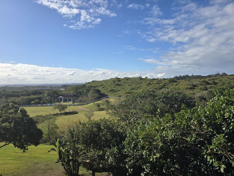 A scenic elevated view overlooking a lush green landscape, rolling hills, and distant estate grounds under a clear blue sky.