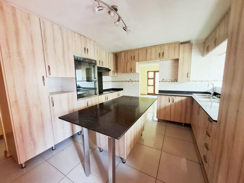 A spacious kitchen featuring light wood cabinetry, dark granite countertops, a central island, built-in oven, and tiled flooring.