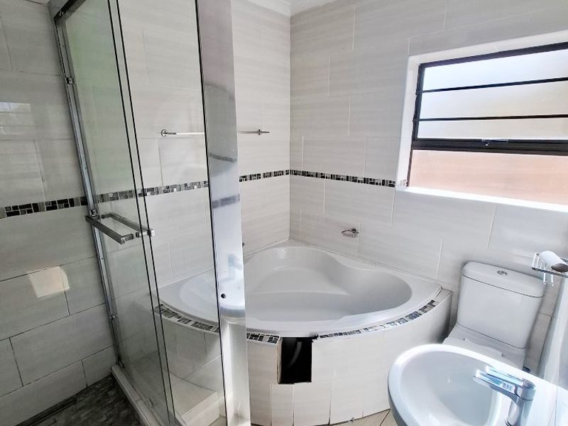 A functional bathroom featuring a corner bathtub, glass-enclosed shower, pedestal basin, and toilet with neutral wall and floor tiling.