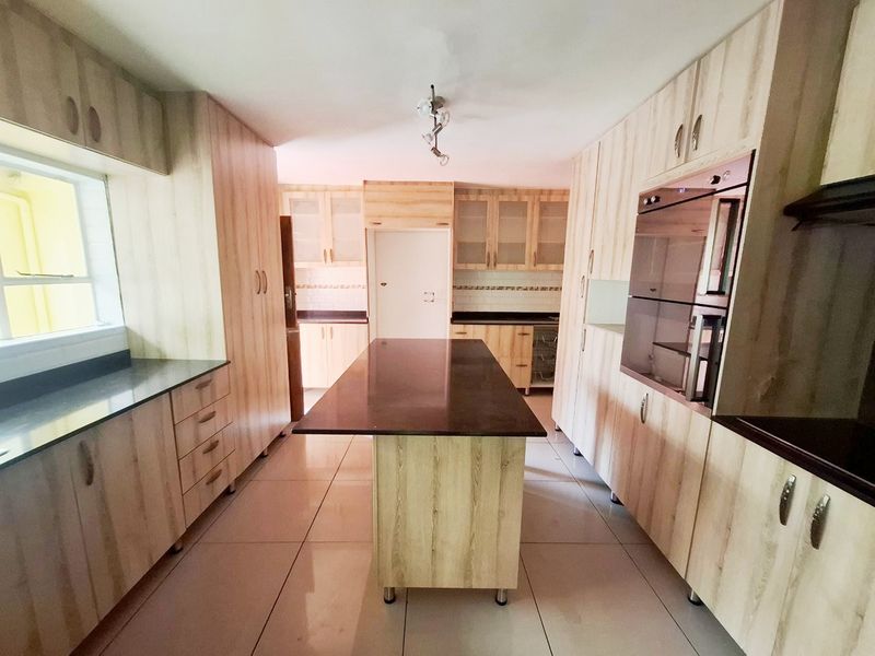 A spacious, modern kitchen featuring light wood-grain cabinetry, dark stone countertops, a central island, and built-in oven, finished with neutral floor tiles.