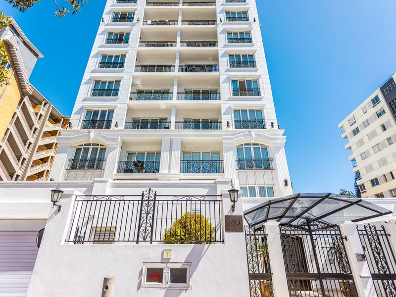 Front elevation of a multi-story apartment building featuring white facade, wrought iron balcony railings, and a secure gated entrance with glass canopy.