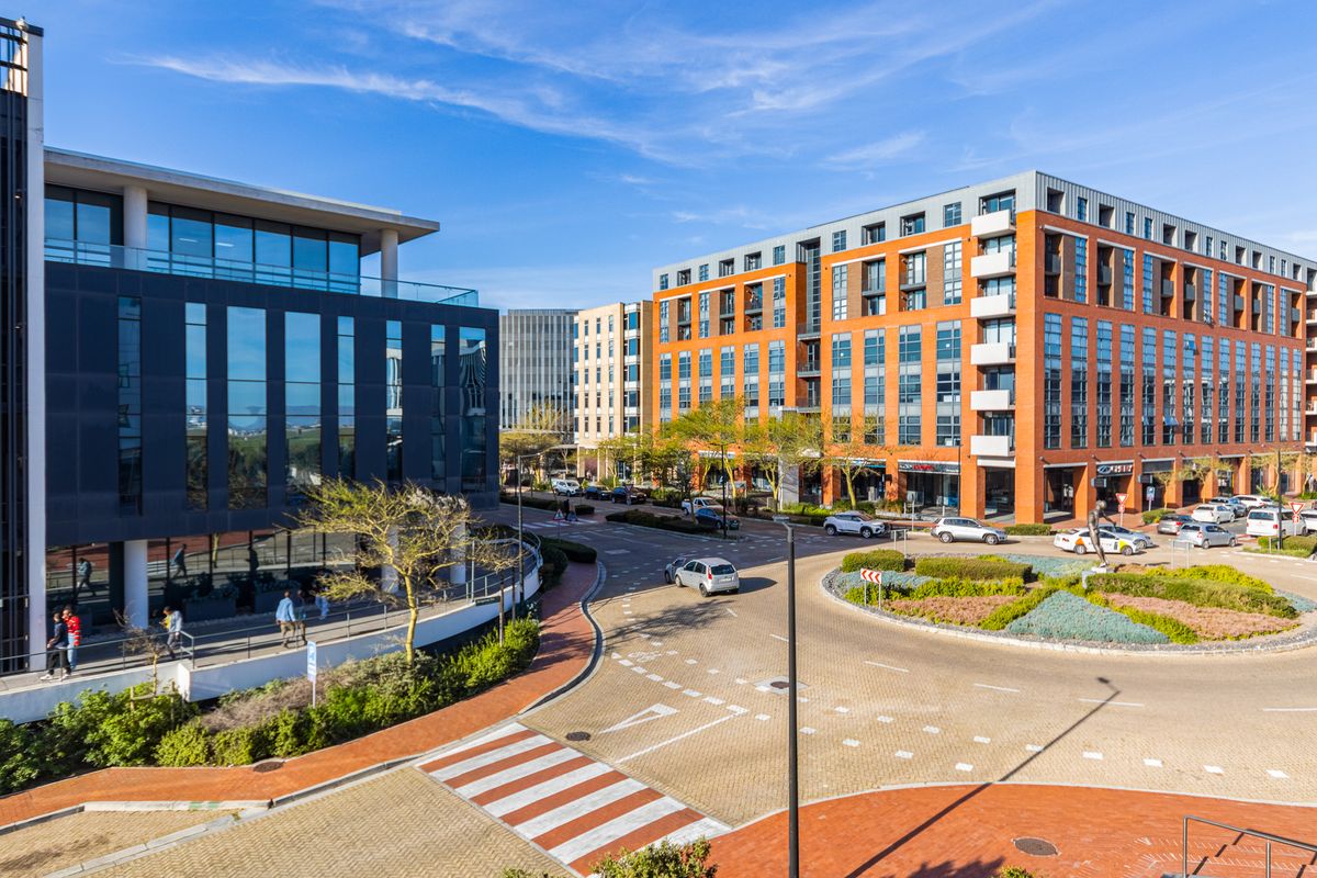 An elevated aerial view of a modern urban precinct featuring multi-story commercial buildings, a paved roundabout, and landscaped public spaces.
