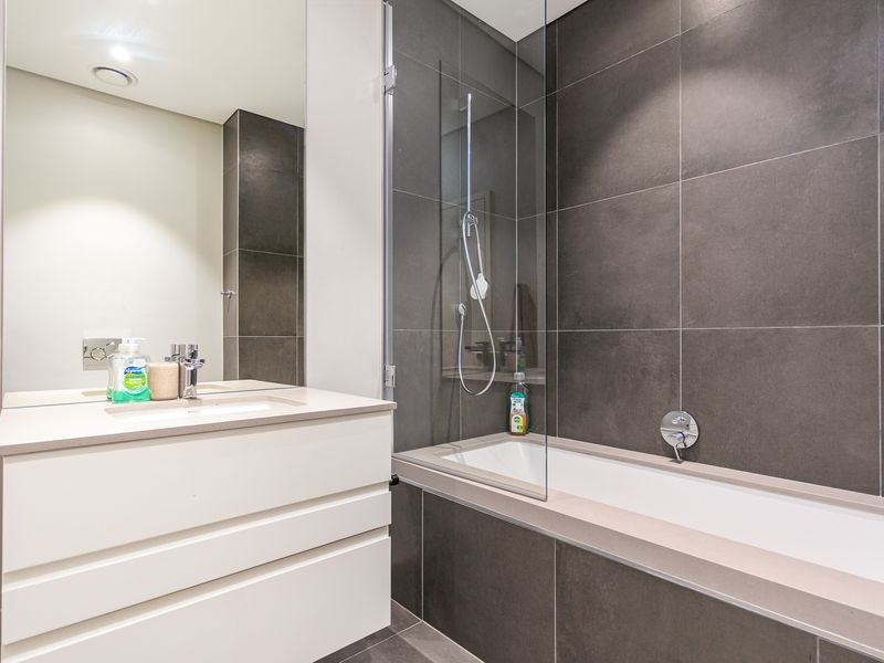 Modern bathroom featuring a sleek white vanity, integrated sink, large mirror, and a bathtub with a glass shower screen and dark tiled finishes.