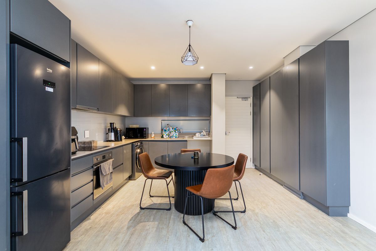 Modern kitchen featuring sleek dark grey cabinetry, integrated appliances, and a central dining area with contemporary lighting and light-toned flooring.