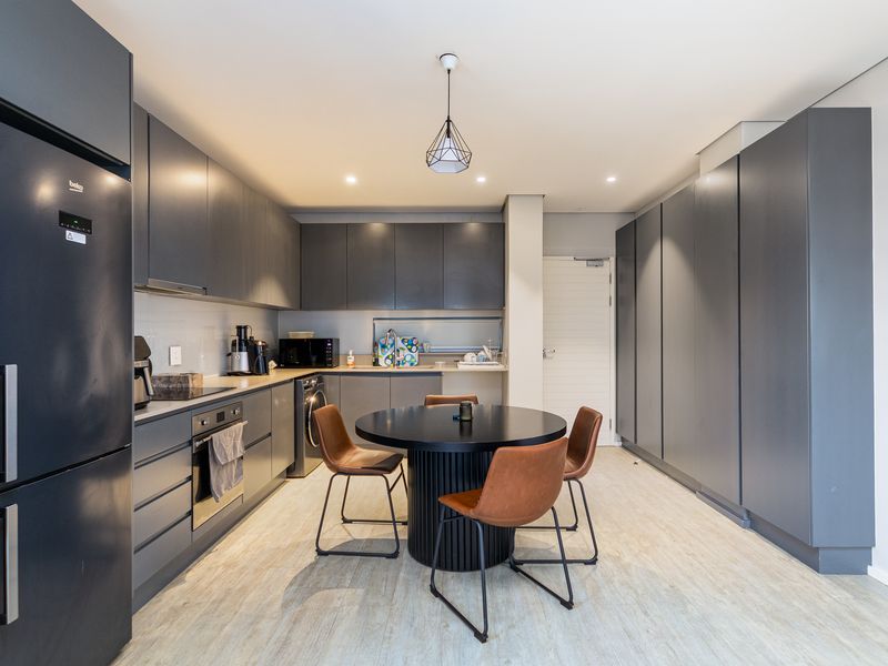 Modern kitchen featuring sleek dark grey cabinetry, integrated appliances, and a central dining area with contemporary lighting and light-toned flooring.