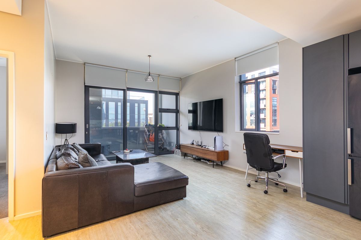 Modern open-plan living area featuring light laminate flooring, large windows with roller blinds, and access to a private balcony.