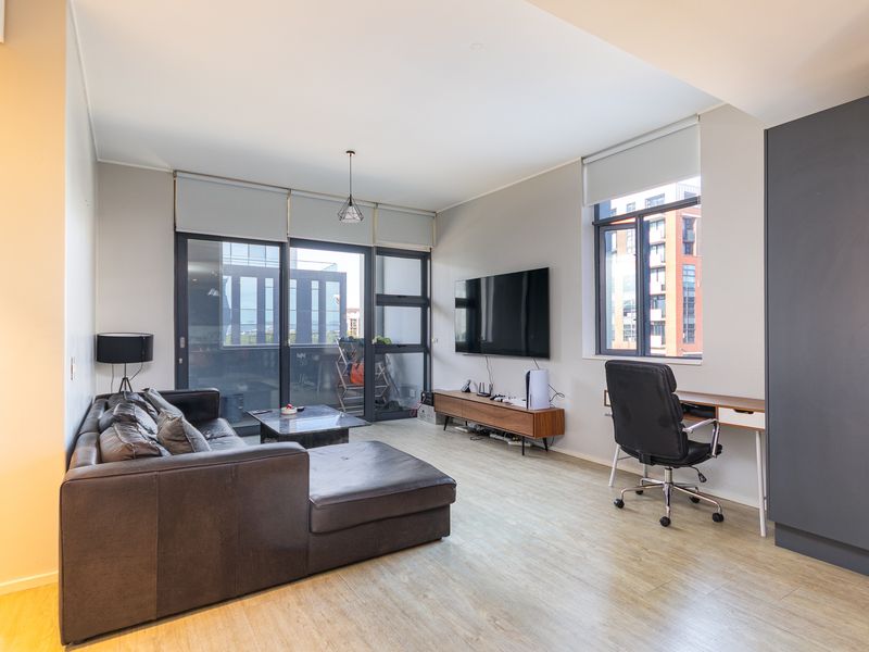 Modern open-plan living area featuring light laminate flooring, large windows with roller blinds, and access to a private balcony.