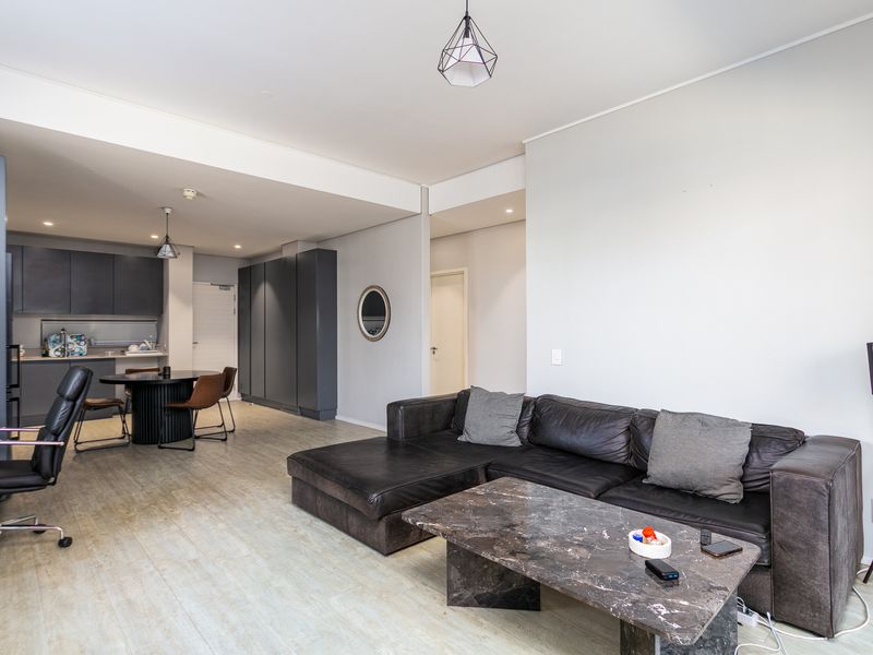 An open-plan living and kitchen area featuring modern dark cabinetry, neutral walls, and light-toned laminate flooring with integrated lighting fixtures.