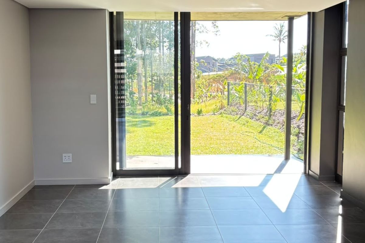 A bright, modern, and empty living area featuring large format floor tiles, recessed lighting, and expansive sliding glass doors opening to a garden view.