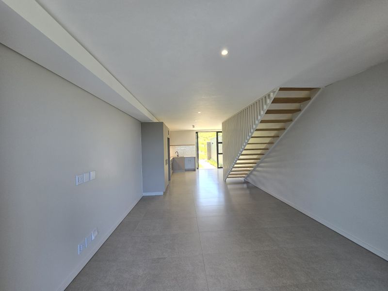 A bright, modern open-plan living area featuring large format floor tiles, a contemporary staircase, and a sleek kitchen leading to an outdoor space.