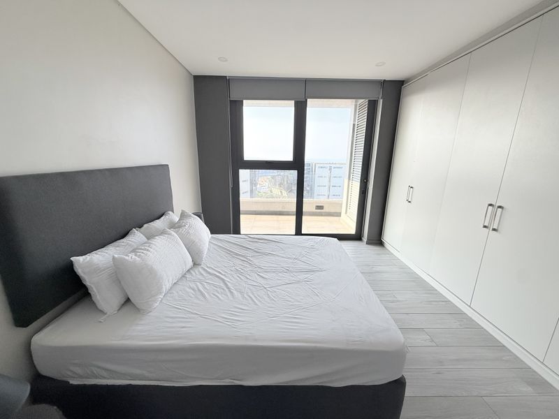 Modern bedroom featuring built-in wardrobes, light laminate flooring, and a large sliding glass door providing access to a balcony with city views.