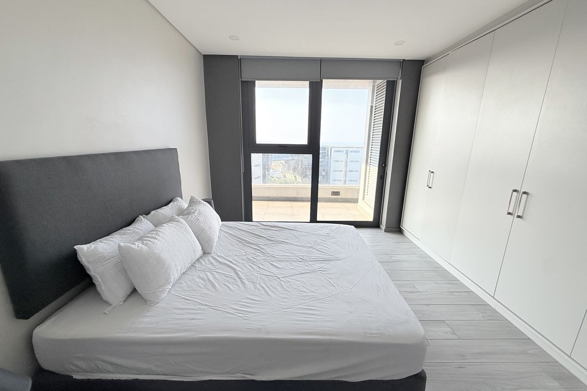 Modern bedroom featuring built-in wardrobes, light laminate flooring, and a large sliding glass door providing access to a balcony with city views.