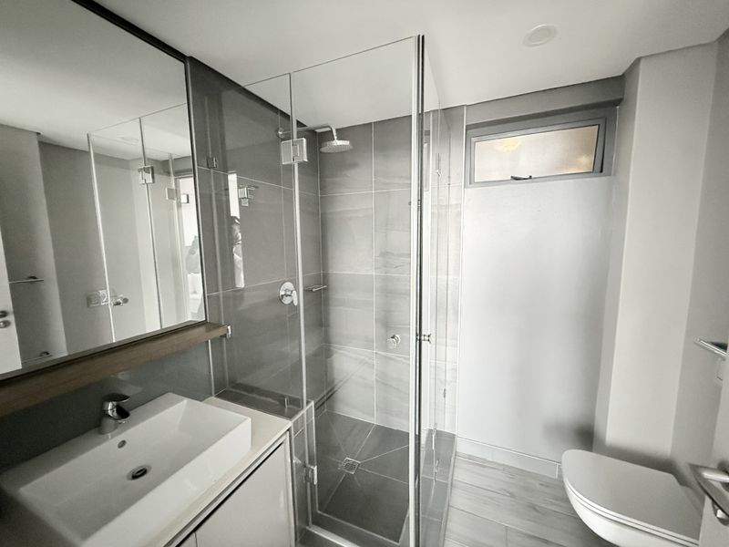 Modern bathroom featuring a glass-enclosed shower, vanity basin, wall-mounted mirror, and contemporary grey tiling throughout.