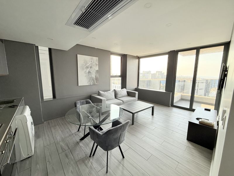 Modern open-plan living area featuring wood-look tile flooring, ducted air conditioning, and large sliding glass doors leading to a balcony with city views.