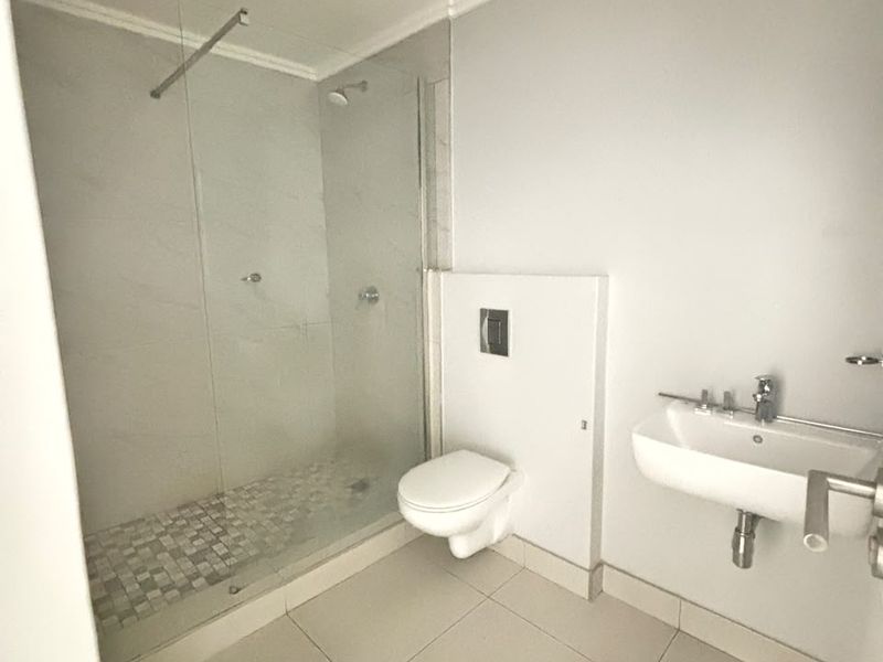 A modern, clean bathroom featuring a walk-in shower with glass screen, wall-mounted toilet, and a wall-mounted basin with neutral floor tiling.