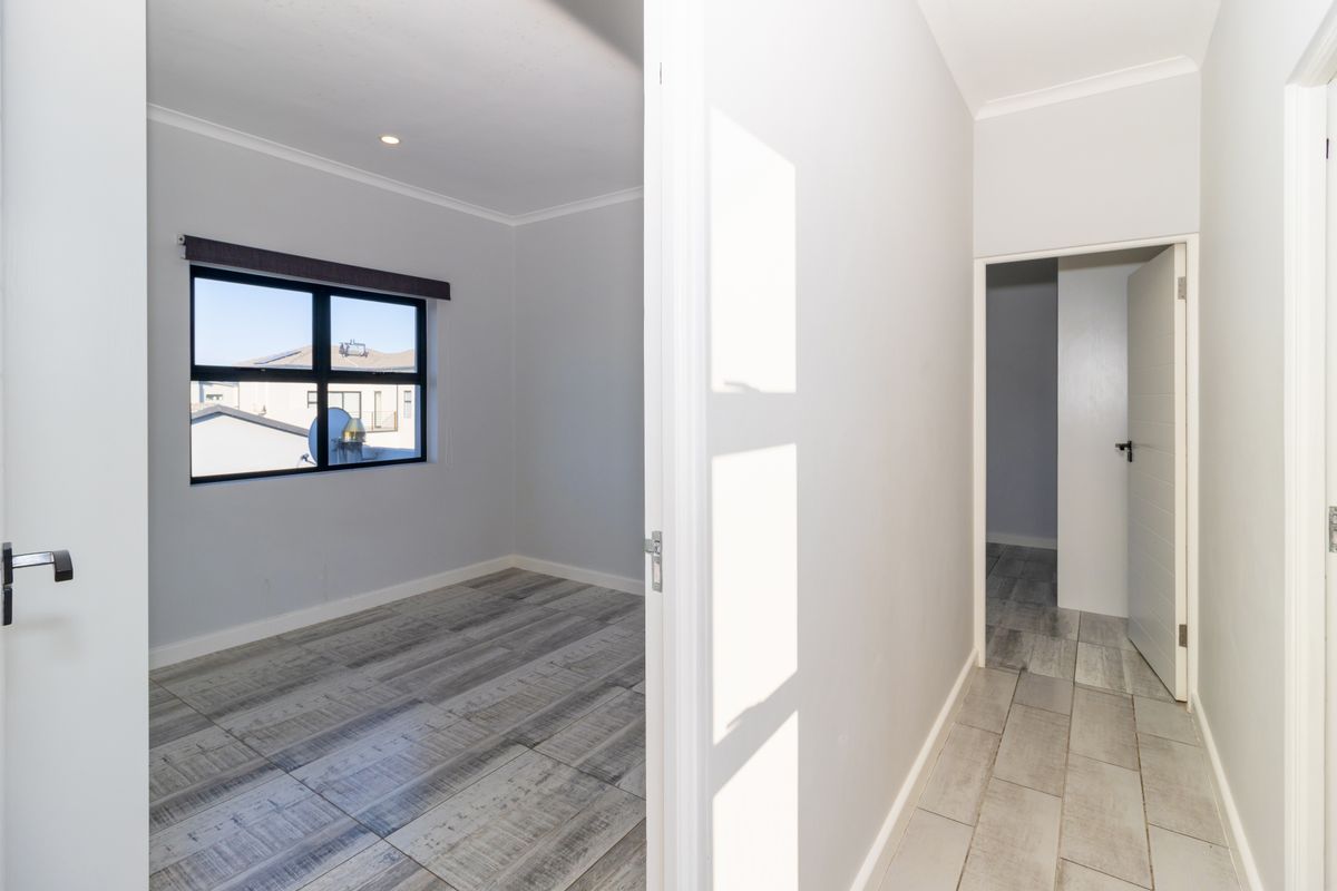 A bright, empty bedroom featuring modern wood-look tiled flooring, neutral walls, and an aluminium-framed window, viewed from an adjacent hallway.