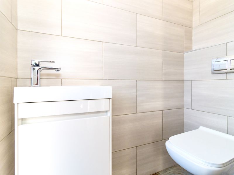 A modern, compact bathroom featuring neutral ceramic wall tiles, a wall-mounted toilet with a concealed cistern, and a sleek vanity basin with chrome fixtures.