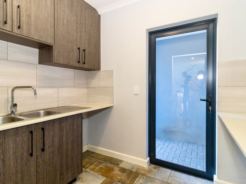 A clean and modern scullery featuring wood-grain cabinetry, stone-look countertops, a stainless steel sink, and direct access to an outdoor paved area.