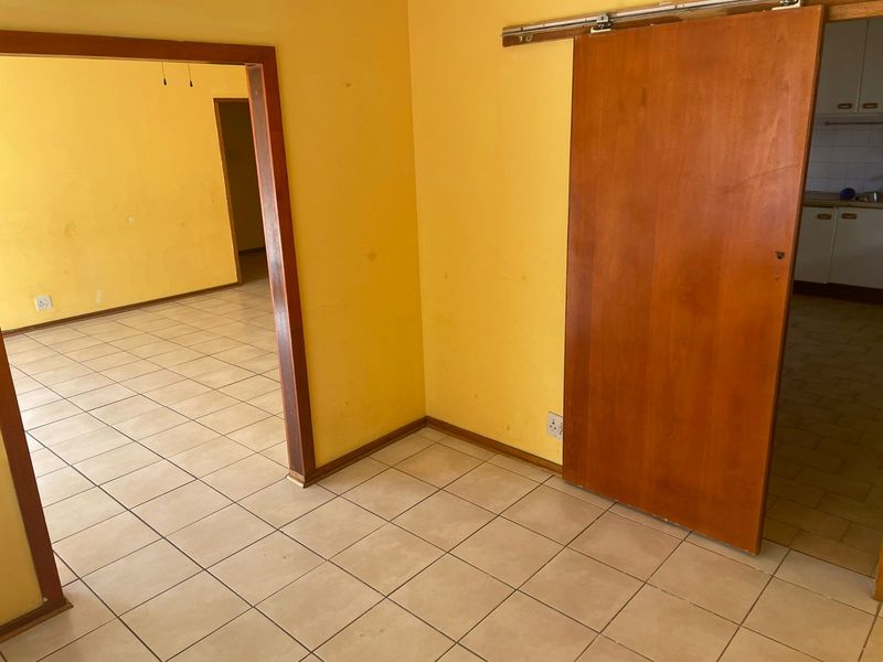 An empty open-plan living area featuring ceramic floor tiles, a sliding wooden door, and yellow painted walls in need of refreshing.