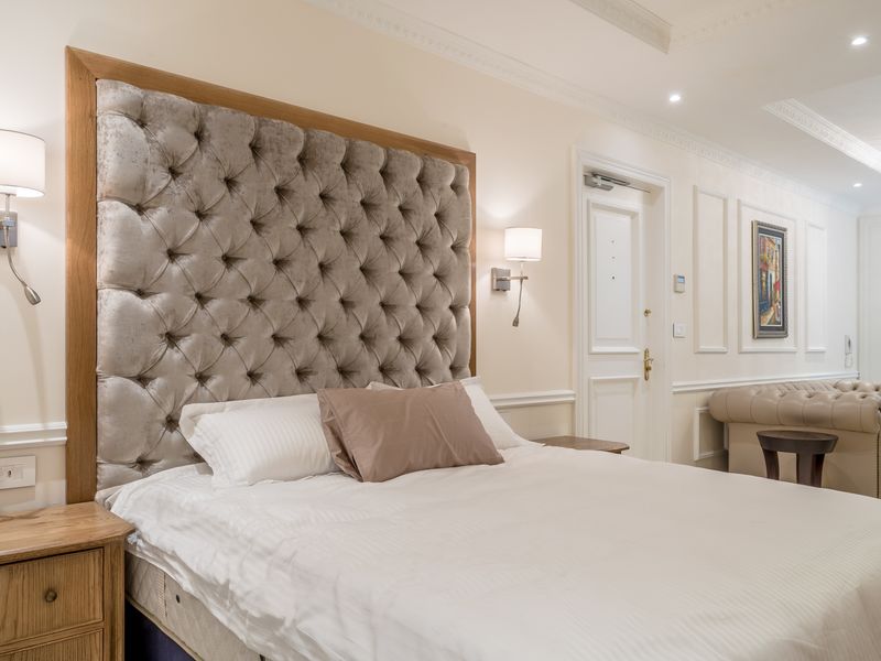 A well-appointed bedroom featuring elegant wall paneling, crown molding, recessed lighting, and integrated wall-mounted fixtures in a neutral, sophisticated color palette.
