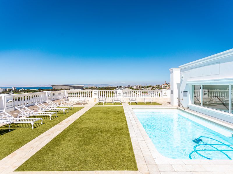 A well-maintained rooftop pool area featuring artificial turf, stone paving, and a classic balustrade with panoramic city and ocean views.