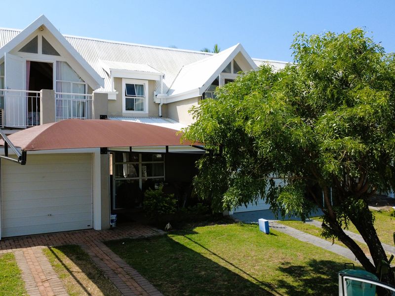 Front elevation of a residential house featuring a pitched metal roof, gabled dormer windows, a balcony, and a paved driveway with a shade sail.