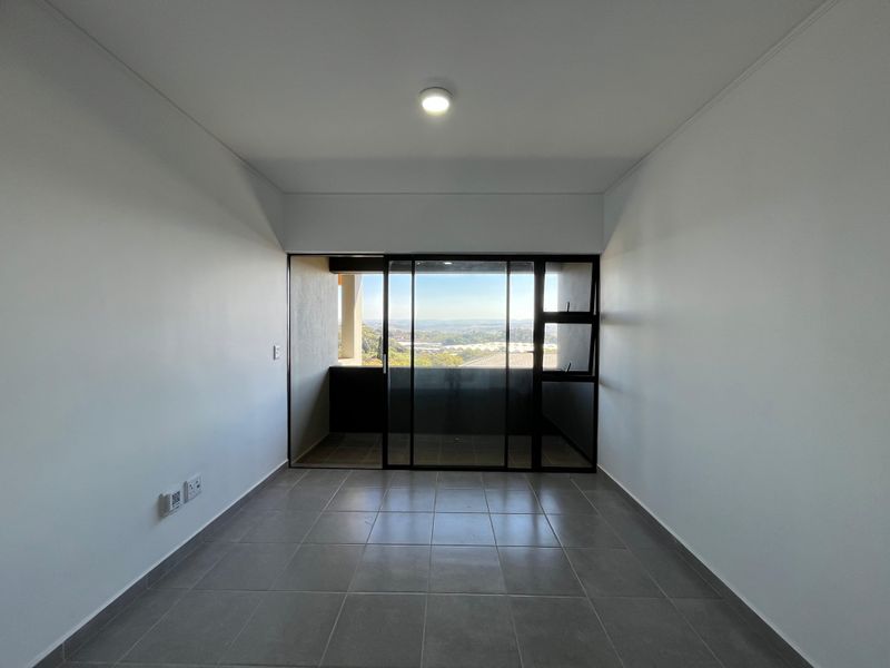 An empty, well-lit living area featuring neutral tiled flooring and a large sliding glass door that opens onto a balcony with scenic views.