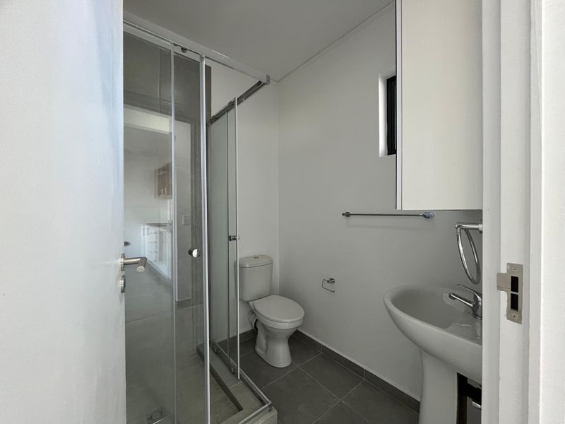 A clean and functional bathroom featuring a pedestal washbasin, a glass-enclosed shower, and a toilet, finished with neutral floor tiles.