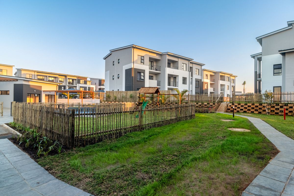 Exterior view of a modern apartment complex featuring a communal garden area, paved walkways, and secure wooden perimeter fencing.