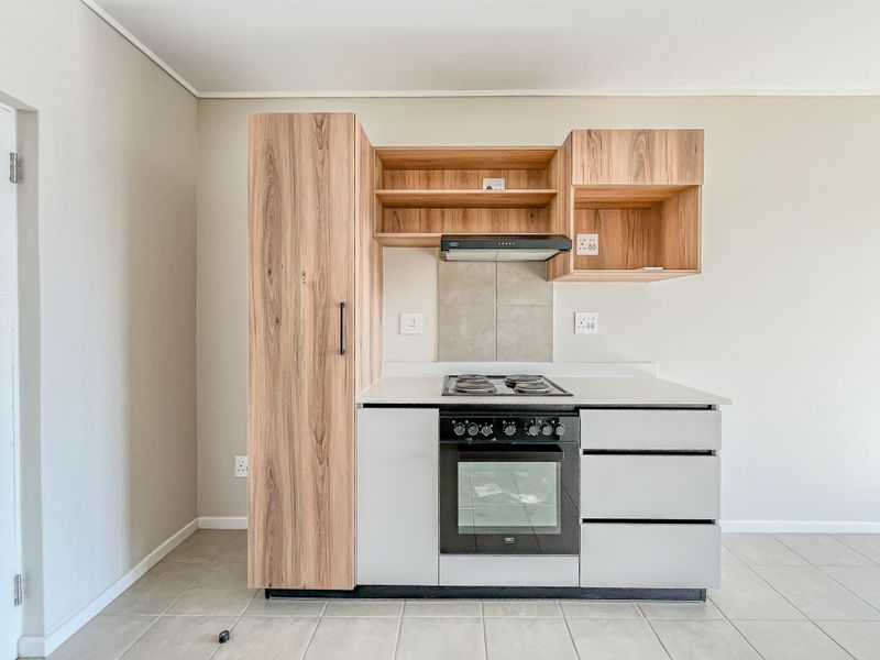 A compact, modern kitchen featuring wood-grain and matte grey cabinetry, a built-in electric oven and hob, and neutral tiled flooring.