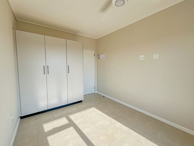 A bright, empty bedroom featuring neutral tiled flooring, a built-in wardrobe, and a ceiling fan, offering a clean and versatile living space.