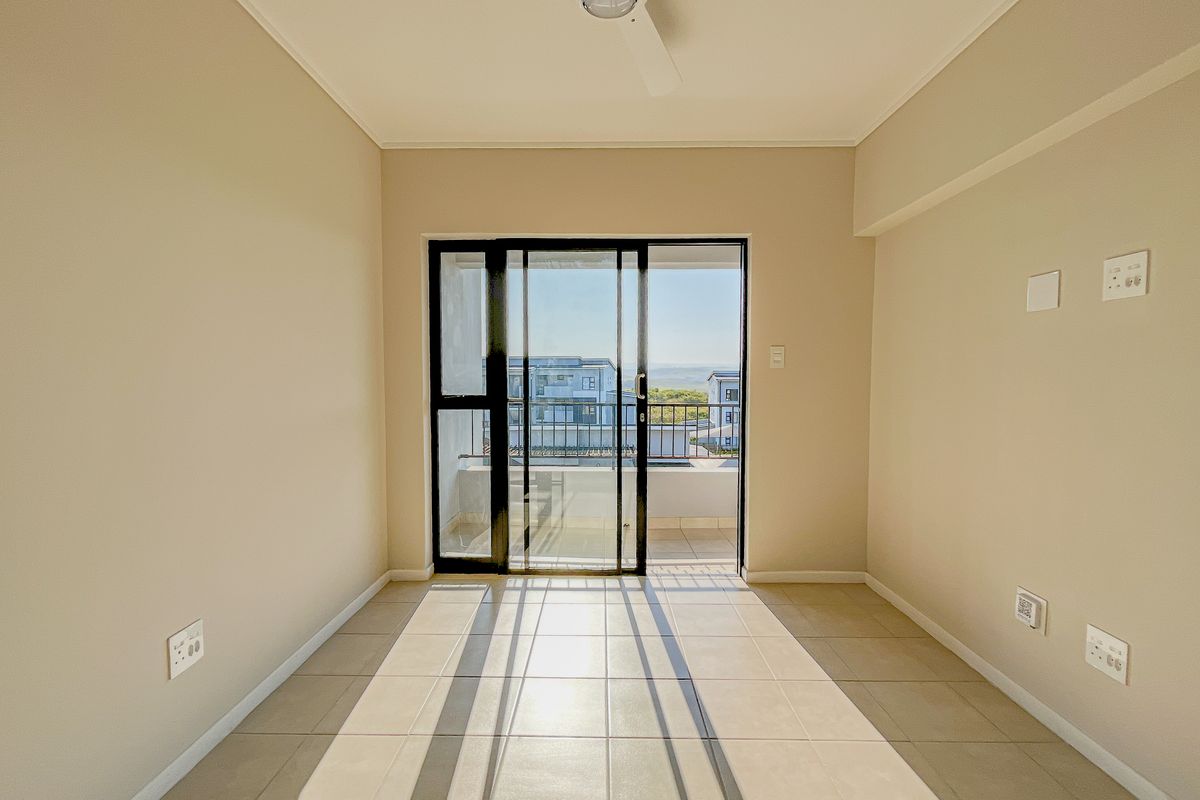 An empty, well-lit living room featuring neutral tiled flooring, a ceiling fan, and a sliding glass door providing access to an outdoor balcony.