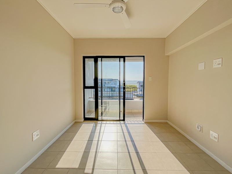 An empty, well-lit living room featuring neutral tiled flooring, a ceiling fan, and a sliding glass door providing access to an outdoor balcony.