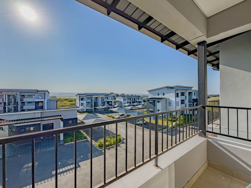 A covered balcony with a metal balustrade, offering an elevated view of the apartment complex and parking area.