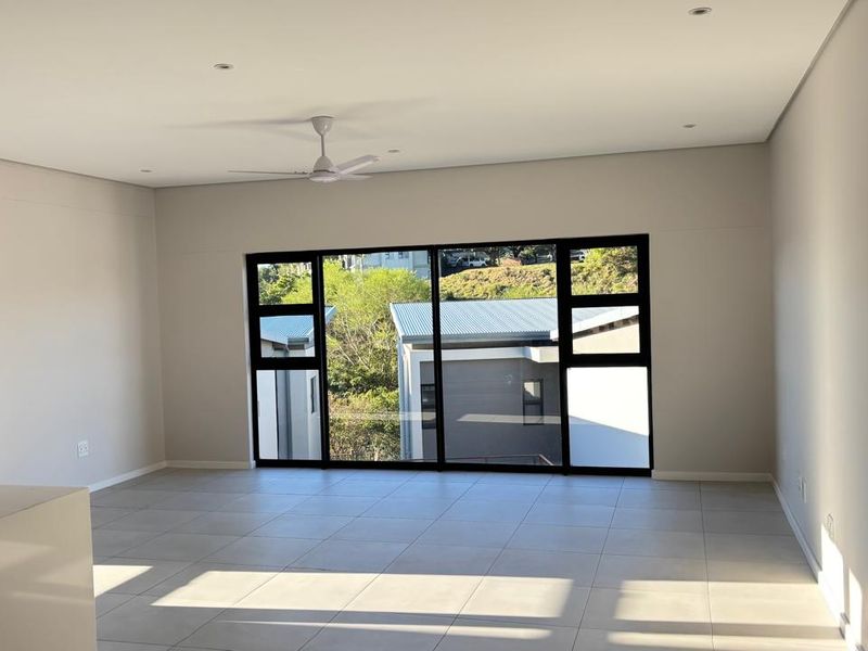 Spacious, light-filled open-plan living area featuring modern tiled flooring, recessed lighting, a ceiling fan, and large windows with views of the surrounding townhouse complex.