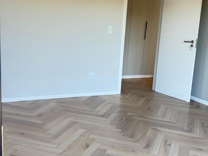 A bright, empty bedroom featuring modern herringbone timber flooring, neutral wall tones, and built-in cupboard storage.