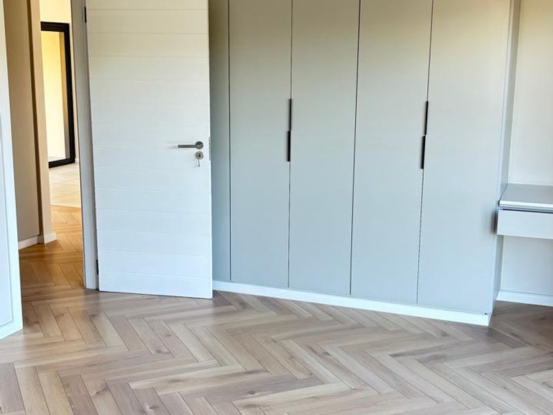 A bright, modern bedroom featuring high-quality herringbone timber flooring and extensive built-in cabinetry with a neutral finish.